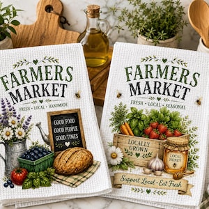 May include: Two white waffle-weave kitchen towels featuring Farmers Market graphics. The left towel shows a metal pitcher with flowers, blueberries, a tomato, bread, and a sign reading Good Food Good People Good Times. The right towel shows produce, honey, and the text Support Local Eat Fresh.