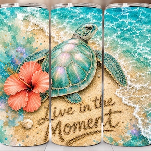 May include: A cylindrical tumbler featuring a watercolour-style beach scene with a sea turtle, a large coral hibiscus flower, and a small seashell on sandy shore. The text "Live in the Moment" is written in a cursive script across the sand.