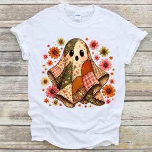 Könnte beinhalten: Ein weißes kurzärmeliges T-Shirt mit einer zentralen Grafik eines Geistes, der aus verschiedenen Patchwork-Stoffmustern in Braun-, Grün- und Orangetönen besteht, umgeben von einer Ansammlung kleiner, bunter Blumen in Rosa, Gelb und Orange.