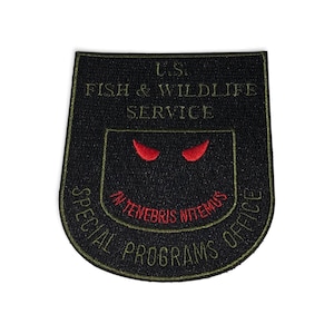 Può includere: Una toppa ricamata nera a forma di scudo con il testo U.S. FISH & WILDLIFE SERVICE in verde oliva, due corna da diavolo rosse sopra il testo rosso IN TENEBRIS NITEMUS e il testo verde oliva SPECIAL PROGRAMS OFFICE in basso.