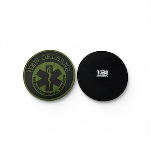 May include: Two circular patches. The left patch is olive green with a black Star of Life symbol and the text NEW ORLEANS EMERGENCY MEDICAL SERVICES. The right patch is solid black with a small white rectangular label reading 13 FIFTY APPAREL.