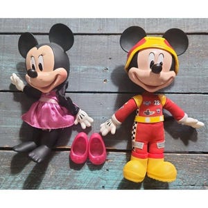 May include: Two plastic character figures. One figure has black ears, a pink dress, and black shoes. The other figure wears a yellow helmet, a red racing suit with the number 28, and yellow boots. Two separate pink shoes are placed between them.