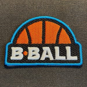 May include: An embroidered patch featuring a stylised orange basketball graphic in the upper arch section, set against a black background. The white text B-BALL is stitched across the lower portion, all bordered by a bright blue thread outline.