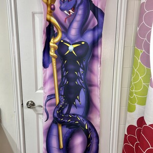 May include: A purple snake-like character with red eyes and a golden staff is depicted on a long, rectangular fabric. The character is wearing a dark outfit with a jewel. The background is a gradient of pink and purple.