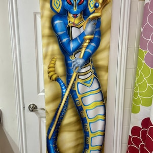 May include: A blue and gold decorative tapestry featuring a cobra-like figure holding a golden staff. The figure has red eyes and is set against a tan background. The tapestry is hanging on a door.