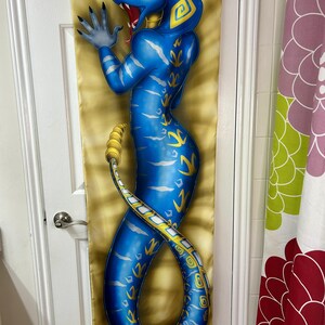 May include: A blue snake-like figure with yellow accents is depicted on a long, rectangular fabric. The figure has a cobra-like head with red accents and is posed with one hand raised. The background is a golden color with a textured pattern.