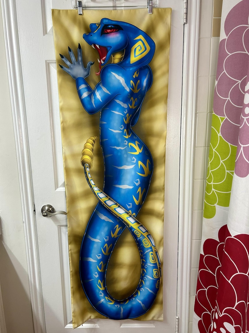 May include: A blue snake-like figure with yellow accents is depicted on a long, rectangular fabric. The figure has a cobra-like head with red accents and is posed with one hand raised. The background is a golden color with a textured pattern.