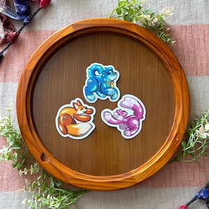 Pode incluir: Três adesivos de vinil cortados com animais de desenho animado projetados como esculturas de balão. Um adesivo é um cão azul, um é uma raposa laranja e um é um gato rosa. Cada adesivo tem um acabamento brilhante com destaques brancos.