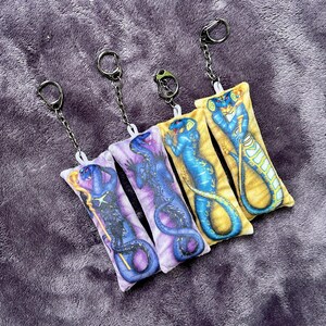 May include: Four rectangular keychains with a snake design in various colors: purple, blue, and yellow. Each keychain has a silver chain and ring. The keychains are lying on a purple, textured surface.