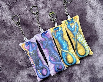 May include: Four rectangular keychains with a snake design in various colors: purple, blue, and yellow. Each keychain has a silver chain and ring. The keychains are lying on a purple, textured surface.