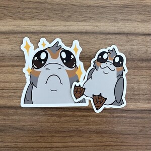 May include: Two stickers featuring cartoon Porgs. The Porgs have large black eyes, gray and brown feathers, and white bodies. One Porg has a starburst effect around it. The stickers are on a wooden surface.