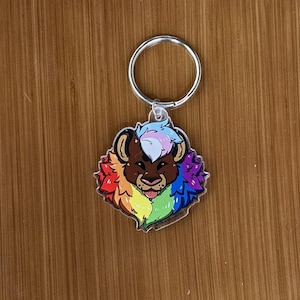 May include: A colorful lion keychain with a rainbow mane and accents. The lion's face is brown with a pink tongue sticking out. The keychain has a silver ring attached to the top.