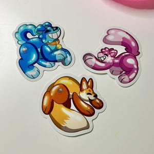 May include: Three cartoon animal stickers: a blue dog with a gold collar, a pink cat, and a brown and orange fox. Each sticker has a glossy, gel-like appearance and a smiling expression. The stickers are on a white surface.
