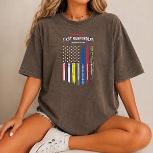 Może przedstawiać: Ciemnoszary T-shirt z krótkim rękawem z grafiką flagi amerykańskiej z pionowymi pasami reprezentującymi służby ratunkowe. Tekst brzmi: America's FIRST RESPONDERS BRAVERY IN ACTION, z etykietami NURSE, EMS, DISPATCH, CORRECTIONS, LAW ENFORCEMENT, FIREFIGHTER oraz MILITARY.