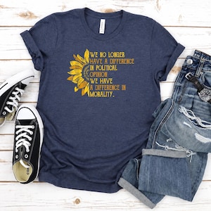 May include: A dark blue short-sleeved t-shirt featuring a yellow sunflower graphic on the left side. The text printed in yellow reads: WE NO LONGER HAVE A DIFFERENCE IN POLITICAL OPINION WE HAVE A DIFFERENCE IN MORALITY. The shirt has a crew neckline.