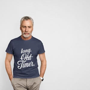 May include: A navy blue short-sleeved t-shirt featuring the white text "long. Old Timer." printed on the chest. The shirt has a crew neckline and a standard fit, providing a clean and simple look for casual wear in various settings.