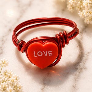May include: A red wire ring with a heart-shaped centerpiece. The heart is red with the word "LOVE" in white. The ring is on a marble surface, with white flowers in the background.