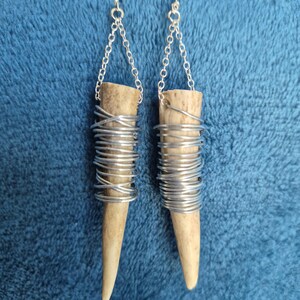 Puede incluir: Un par de pendientes colgantes con segmentos de asta natural de color marrón claro, envueltos en alambre de tono plateado. Cada pendiente cuelga de una cadena de metal plateado y un gancho estándar, creando un diseño largo y texturizado.