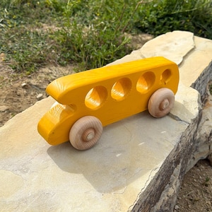 May include: A bright yellow wooden toy school bus with four wooden wheels. The bus has four circular windows and is sitting on a large, light-colored rock. The background features green grass and foliage.