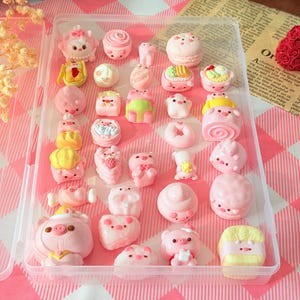 May include: A clear plastic storage container filled with an assortment of small, pink, pig-themed miniature figurines and food-shaped charms. The collection includes various rounded pig faces, pig-shaped pastries, and small decorative items.