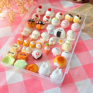 May include: A clear plastic storage container filled with an assortment of miniature resin figures, including various white seals, food-shaped items, and colorful characters. The text Kawaii Seal is printed in white on a light pink rectangular background.