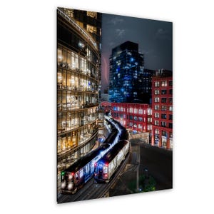 May include: A night cityscape featuring a train traveling on elevated tracks between a curved glass-walled building and red brick structures. The scene includes illuminated windows, a dark sky with a faint lightning bolt, and city lights.