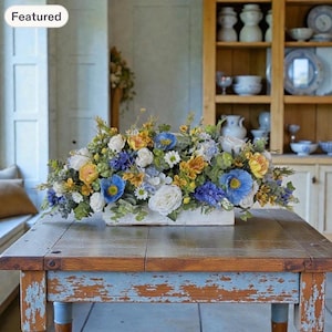 May include: A floral centerpiece arrangement featuring blue, yellow, and white flowers with green foliage in a rectangular white container. The arrangement includes poppies, roses, and small daisies, creating a dense, multi-colored display.