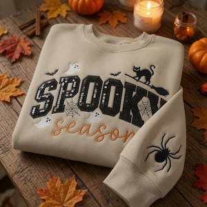 May include: A beige crewneck sweatshirt featuring the word SPOOKY in black sequined block letters and the word season in orange cursive embroidery. The design includes embroidered white ghosts, black bats, a black cat on a broom, and a spider.