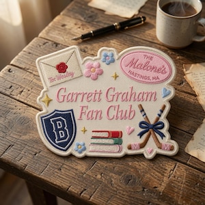 May include: An embroidered patch featuring the text Garrett Graham Fan Club in pink, with icons including an envelope with a red wax seal, a blue shield with a B, a stack of books, and crossed hockey sticks with a blue bow on a cream background.