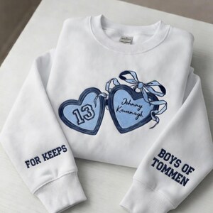 May include: White sweatshirt with blue embroidered heart-shaped patches. One heart has the number 13, the other says "Johnny Kavanagh". The sleeves read "FOR KEEPS" and "BOYS OF TOMMEN" in blue. A light blue bow is between the hearts.