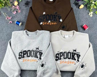 May include: Three crewneck sweatshirts in brown, light gray, and cream, each featuring the text SPOOKY season in black and orange, accompanied by small ghost and cat graphics, with a black spider illustration on the left sleeve.