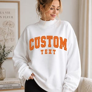 May include: A white long-sleeve sweatshirt with a mock neck collar. The front features the word CUSTOM in large, orange, polka-dotted block letters, with the word TEXT printed in smaller, solid orange block letters directly underneath.