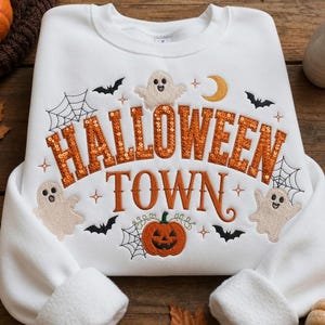 May include: A white long-sleeve sweatshirt featuring the embroidered text HALLOWEEN TOWN in orange sequins and brown thread. The design includes three white ghosts, black bats, spider webs, a yellow crescent moon, and an orange pumpkin.