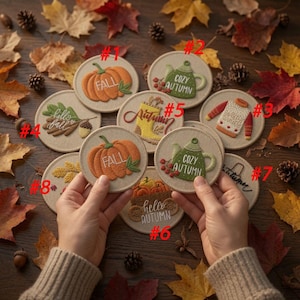 May include: A collection of round fabric coasters featuring embroidered autumn designs including pumpkins, teapots with the text 'COZY AUTUMN', sweaters, and boots. Each coaster is numbered from 1 to 8, showcasing seasonal motifs in warm colors.