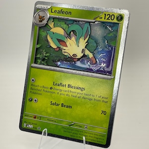 Puede incluir: Una carta coleccionable Pokémon de Leafeon, verde y amarilla. La carta presenta una ilustración de Leafeon, con el texto "Leafeon" y "120 HP". El texto describe las habilidades "Leaflet Blessings" y "Solar Beam".