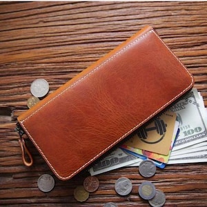 May include: A rectangular brown leather wallet with white stitching and a zipper closure. The wallet is partially open, revealing a stack of one hundred dollar bills and several credit cards. Silver coins are scattered around the wallet.
