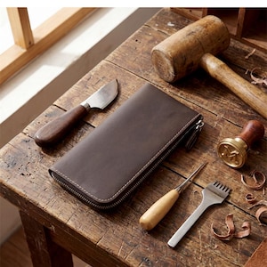 Könnte beinhalten: Eine dunkelbraune Ledergeldbörse mit Reißverschluss und weißen Nähten, begleitet von einem Holzhammer, einem gebogenen Ledermesser, einer Metallahle und einem Stechbeitel mit dem Buchstaben M auf einem Messingstempel.