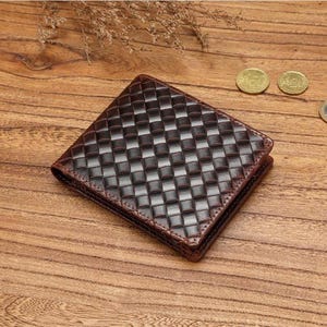 Pode incluir: Uma carteira dobrável castanho-escura com um padrão entrançado em forma de grelha na superfície exterior. A carteira tem uma forma quadrada com cantos arredondados e um acabamento brilhante, exibindo uma textura consistente.
