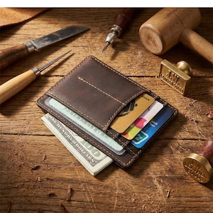 Peut inclure: Un porte-cartes en cuir marron foncé avec des coutures apparentes, contenant plusieurs cartes de crédit et une liasse de billets. Le portefeuille est posé sur une surface en bois avec divers outils de maroquinerie et un tampon en métal.