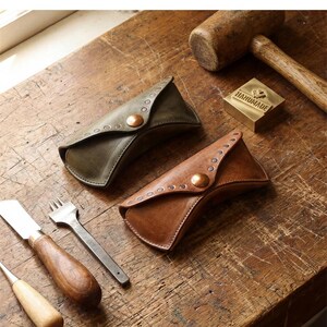 May include: Two leather eyeglass cases, one olive green and one tan, featuring a triangular flap with a central copper-colored snap button and decorative metal studs along the edge. A wooden mallet and metal tools are placed nearby.