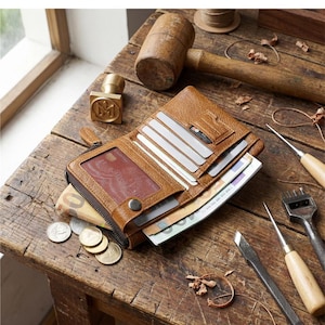 May include: A tan leather bifold wallet with a zippered compartment, multiple card slots, and a clear ID window. The wallet is open, revealing paper currency inside, and is surrounded by various metal and wooden leatherworking tools.