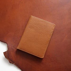 Puede incluir: Un tarjetero o billetera rectangular de cuero color canela con una textura granulada distintiva y costuras a tono a lo largo de los bordes. El artículo presenta un diseño plegado y un color marrón claro uniforme en toda su superficie.