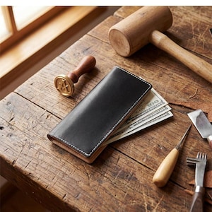 May include: A black leather long wallet with white stitching containing United States hundred-dollar bills. The wallet is positioned near a wooden mallet, a metal stamp with the letter A, a metal awl, and a metal edge creaser tool.