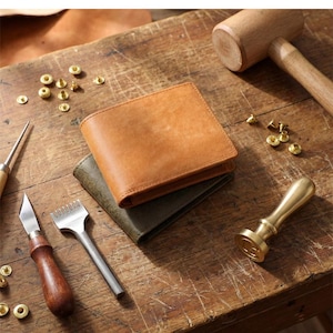 Op de afbeelding: Een beige lederen portemonnee gestapeld op een olijfgroene lederen portemonnee, omringd door metalen klinknagels, een houten hamer, een hobbymes, een stikbeitel en een messing stempel op een houten werkbank.