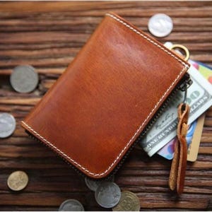 May include: A brown leather wallet with a zipper closure and white stitching. The wallet contains a one hundred dollar bill and a credit card. Several silver coins are scattered around the wallet, and a leather pull tab is attached to the zipper.
