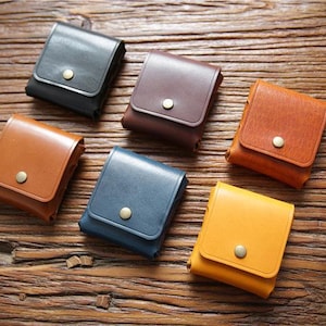 Könnte beinhalten: Sechs kleine quadratische Taschen aus glattem Leder in Schwarz, Dunkelbraun, Hellbraun, Blau, Orange und Gelb. Jede Tasche verfügt über einen Klappenverschluss, der mit einem mittigen Metalldruckknopf gesichert ist und kompakten Stauraum bietet.