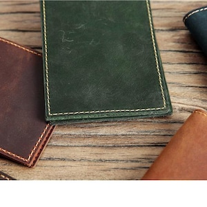 May include: A collection of rectangular leather card holders in various colors including dark green, brown, tan, and black. Each item features visible light-colored stitching along the edges and a smooth, matte finish on the surface of the leather.