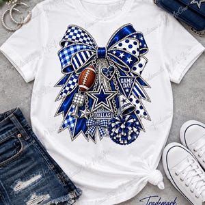 Puede incluir: Una camiseta blanca de manga corta con un gran gráfico de un lazo azul y blanco. El lazo está adornado con dijes que incluyen un balón de fútbol americano, una estrella, un megáfono, un pompón y pancartas que dicen GAME DAY y DALLAS.
