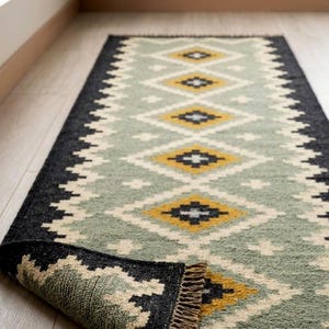 May include: A rectangular runner rug featuring a geometric diamond pattern in sage green, mustard yellow, black, and cream. The rug has a thick black border with a zigzag edge and short, light brown fringe at the end, which is folded over to show the back.
