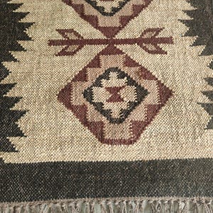 May include: A woven runner rug featuring a textured beige base with dark brown borders and a central pattern of geometric diamond shapes in shades of maroon and black. The rug is finished with a fringe of loose threads along the bottom edge.